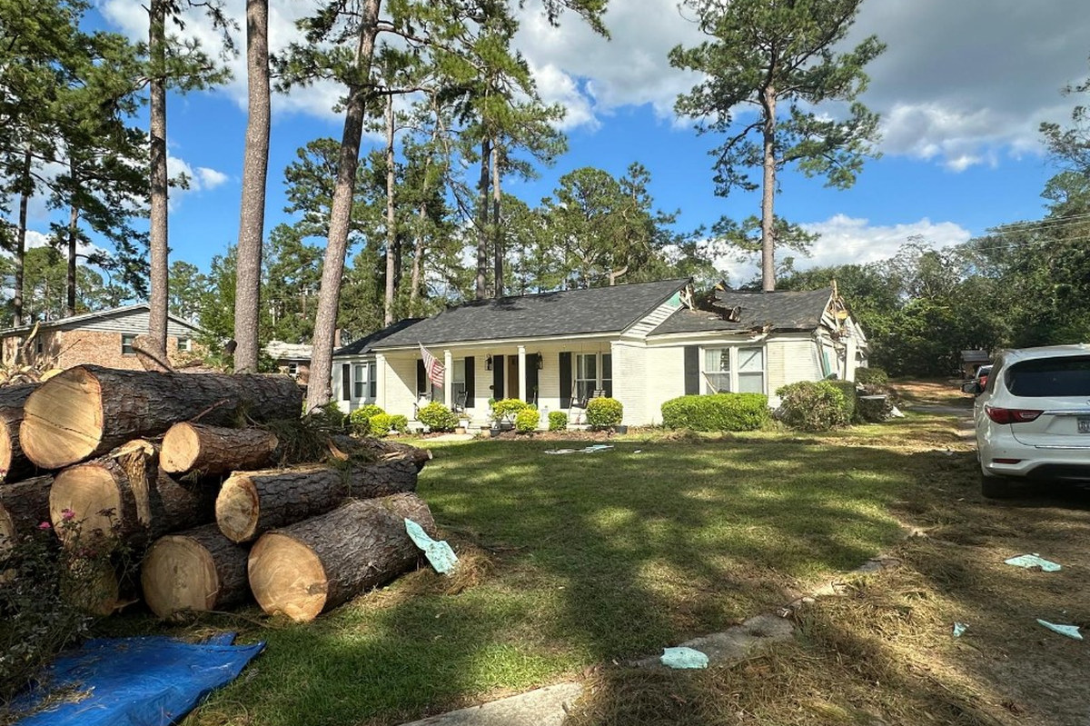 Fallen Tree Roof Damage & Cut Log Cleanup | Warm Home Inc Emergency storm tree removal — stacked cut pine logs in the yard of a white house with a crushed roof after a fallen tree | Warm Home Inc