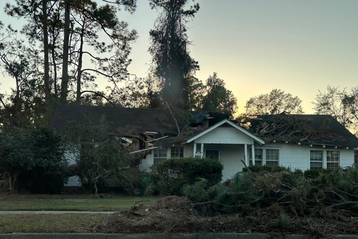 Tree Through Roof Storm Damage Cleanup | Warm Home Inc Emergency tree removal — white house with roof and wall crushed by a fallen tree, storm debris piled on the front lawn | Warm Home Inc